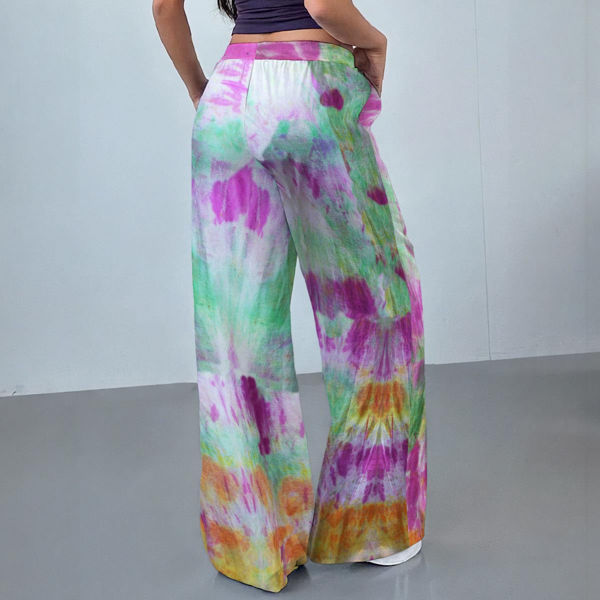 A person wearing wide-leg tie-dye pants, shown from the back in a studio setting. The pants have a colorful spiral tie-dye pattern with green, purple, pink, orange, and white.