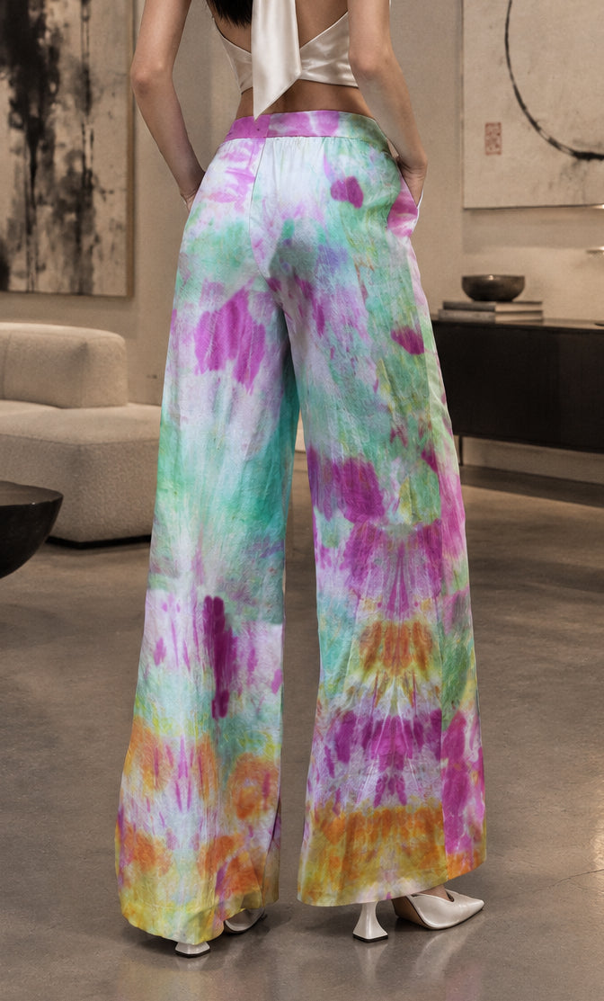 A person wearing wide-leg tie-dye pants, shown from the back in a studio setting. The pants have a colorful spiral tie-dye pattern with green, purple, pink, orange, and white.