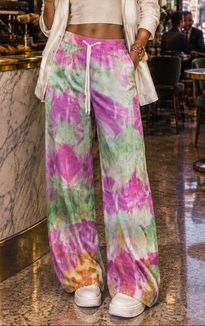 Wide-leg tie-dye sweatpants in a vibrant pink, green, purple, and orange pattern, featuring a white drawstring waist, modeled on a person against a gray studio background.
