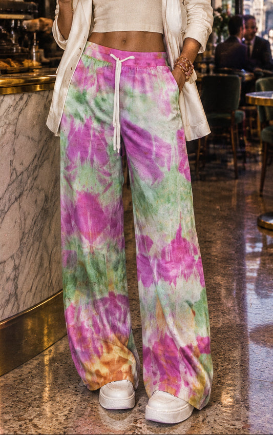 Wide-leg tie-dye sweatpants in a vibrant pink, green, purple, and orange pattern, featuring a white drawstring waist, modeled on a person against a gray studio background.