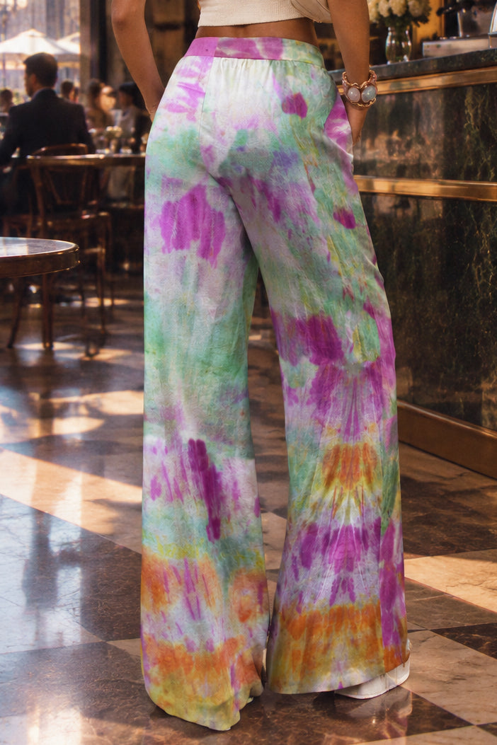 A person wearing wide-leg tie-dye pants, shown from the back in a studio setting. The pants have a colorful spiral tie-dye pattern with green, purple, pink, orange, and white.