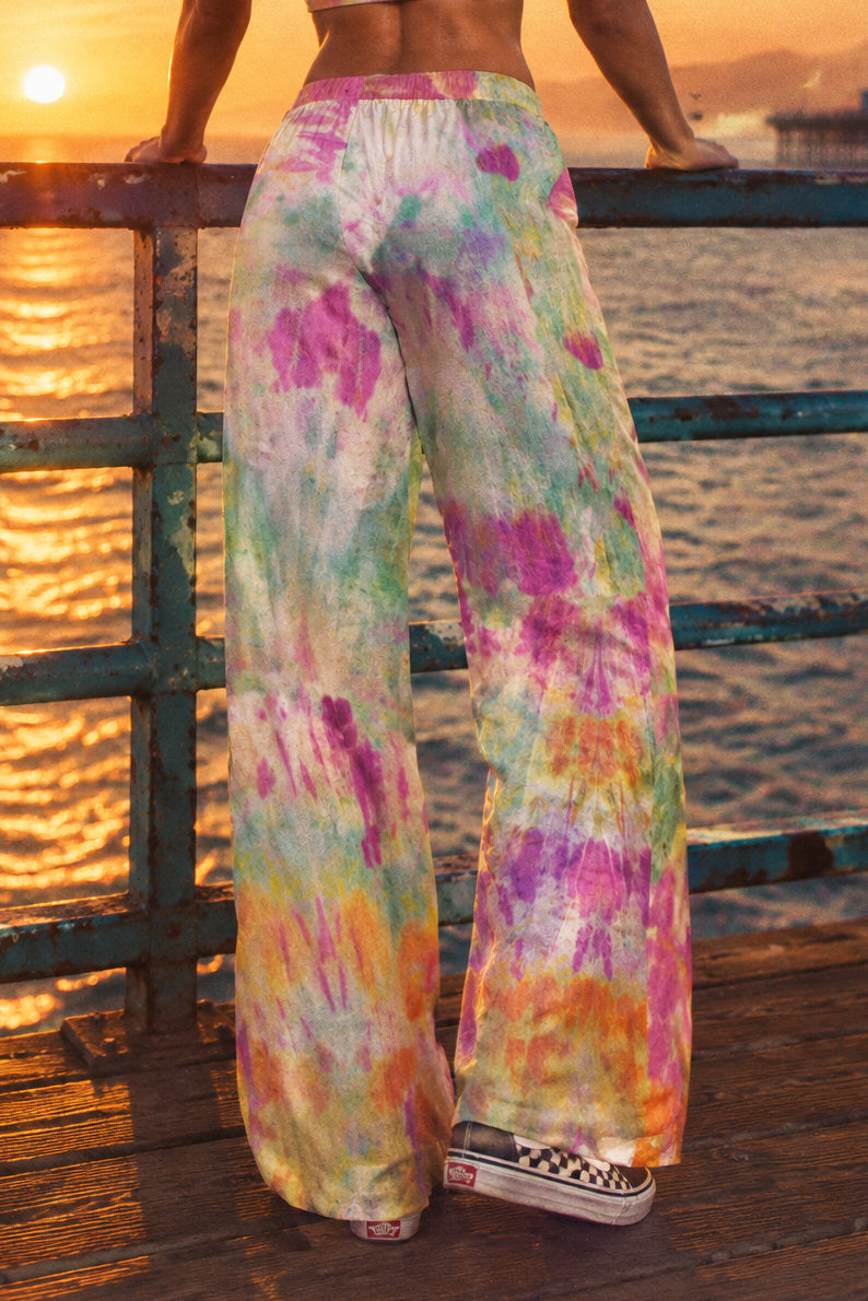 A person wearing wide-leg tie-dye pants, shown from the back in a studio setting. The pants have a colorful spiral tie-dye pattern with green, purple, pink, orange, and white.